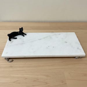 May include: A rectangular white marble serving tray featuring a black cat figurine attached to the top left corner and small silver-toned metal feet shaped like cat paws supporting the base of the tray on the surface.