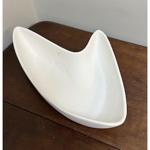 May include: A white serving dish in a mid-century modern style. The dish has an unusual, asymmetrical shape with a curved, wing-like design. The dish is made of a smooth, solid white material and is empty.