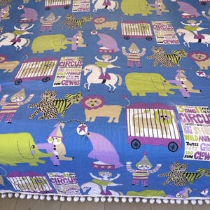 May include: A blue fabric featuring a repeating circus pattern with green elephants, lions in cages, acrobats on horses, clowns, and leopards. The text reads Circus, Big Show, Wild Animals, and Fun Clowns. The bottom edge has a white pom-pom trim.