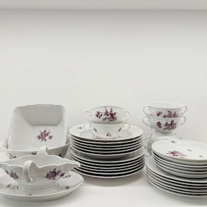 May include: A white porcelain dinnerware set with a purple floral pattern. The set includes plates, bowls, cups, and a serving dish. The plates are stacked in neat piles, and the set is arranged on a white surface.