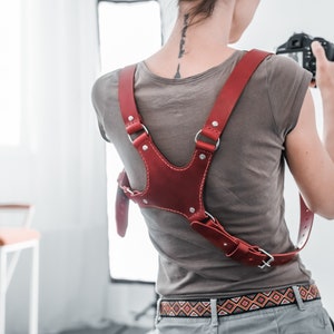 Pode incluir: Uma correia de câmera de couro vermelho com correias ajustáveis e fivelas de metal. A correia é usada sobre os ombros e cruzada sobre o peito.