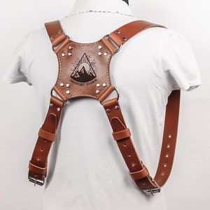 May include: Brown leather camera strap with adjustable buckles and a metal ring. The strap features a mountain design with the text "Enchantment Studios" engraved on it.