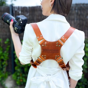 May include: A woman wearing a white blazer with a brown leather camera strap. The strap has the text "MANON BOTERDAEL PHOTOGRAPHIE" on it.