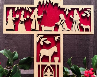 May include: A wooden cross with a red background and a cutout design depicting the Nativity scene. The figures include Mary, Joseph, the baby Jesus, a donkey, a sheep, and three wise men. The cross is decorated with a star and leaves.