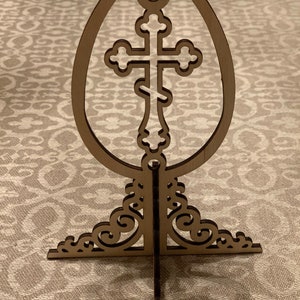 May include: Wooden Easter egg stand with a cross cutout. The stand is made of brown wood and has a delicate, ornate design.