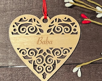 May include: A wooden heart-shaped ornament with a delicate, intricate design and the word "Baba" engraved on it. The ornament is hanging from a red ribbon.