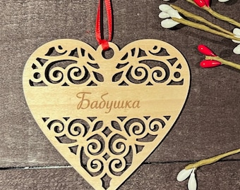 May include: A wooden heart-shaped ornament with intricate cut-out designs and the word "Babushka" in script. It hangs from a red ribbon. The ornament is made of light-colored wood and has a decorative pattern.