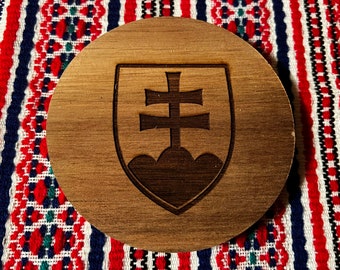 May include: A wooden coaster with a laser-engraved design of the Slovakian coat of arms. The coat of arms features a double cross on a shield with three mountains at the base.