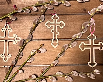 May include: Three wooden cross ornaments with intricate designs, hanging from willow branches with fuzzy buds. The crosses are a light beige color and have a traditional, ornate style.