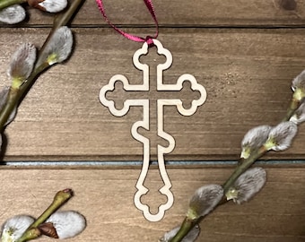 May include: A wooden cutout of a traditional Orthodox cross with a delicate, intricate design. The cross is hanging from a red ribbon and is displayed against a rustic wooden background.