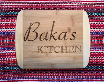 May include: A rectangular bamboo cutting board with rounded corners. The words "Baka's KITCHEN" are engraved in a dark brown font. The board is set against a colorful patterned fabric.