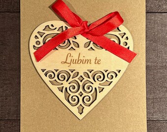 May include: A brown card with a wooden heart cutout with a red ribbon tied around it. The heart has a decorative design and the text "Ljubim te" is written inside.