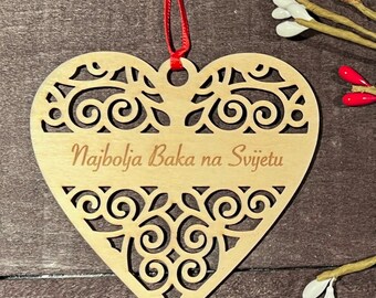 May include: A wooden heart-shaped ornament with a decorative cutout design. The ornament is painted a light brown color and has the text "Najbolja Baka na Svijetu" engraved on it. The ornament is hanging from a red ribbon.