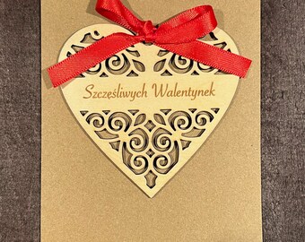 May include: A brown card with a wooden heart cutout with a red ribbon tied around it. The heart has a decorative design and the text "Szczesliwych Walentynek" is written inside the heart.