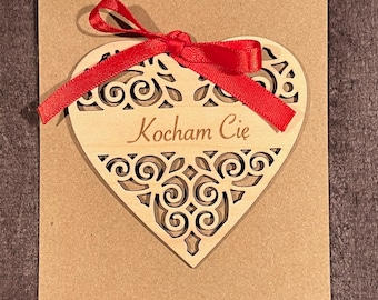 May include: A brown card with a wooden heart cutout attached with a red ribbon. The heart has a decorative design and the text "Kocham Cie" in the center.