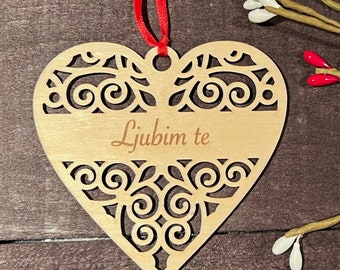 May include: A wooden heart-shaped ornament with a decorative cutout design and the text "Ljubim te" in cursive. The ornament is attached to a red ribbon.