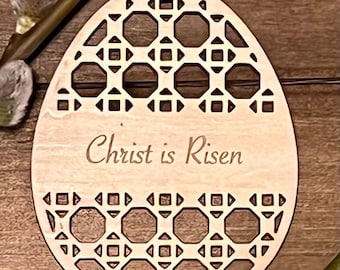 May include: A wooden Easter egg ornament with a geometric pattern and the text "Christ is Risen" engraved on the front. The ornament is light brown and has a red ribbon attached to the top.
