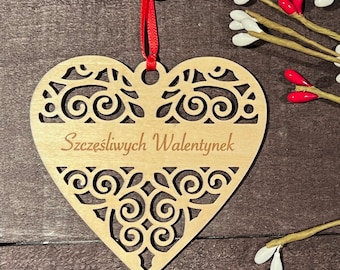 May include: A wooden heart-shaped ornament with intricate cut-out designs and the text "Szczęśliwych Walentynek" in a script font. It hangs from a red ribbon. Decorative branches with white and red accents are in the background.