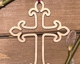 May include: A wooden cross ornament with a decorative design, hanging from a red ribbon. The cross is on a brown wooden background with a branch of willow.