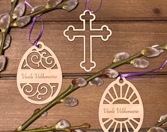 May include: Three wooden ornaments with a spring theme. The ornaments are shaped like a cross and two eggs. The cross has a simple design and the eggs have intricate patterns. The ornaments are painted in a natural wood color and have the text "Vesele Velikonočne" written on them.