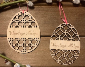 May include: Two wooden Easter egg ornaments with intricate cut-out designs and the text "Wesołego Alleluja". Each egg is suspended by a pink ribbon and set against a wooden background with pussy willow branches.