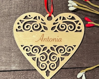 May include: A wooden heart-shaped ornament with a decorative cutout design and the name "Antonia" engraved on it. The ornament is hanging from a red ribbon.