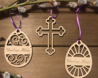 May include: Three wooden Easter ornaments with purple ribbon hangers. The ornaments are a cross, an egg with a sunburst design, and an egg with a floral design. Each ornament has the text "Wesołego Alleluja" which translates to "Happy Easter" in English.