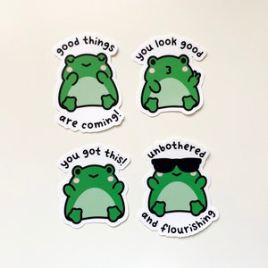 May include: Four frog stickers with different messages. The first sticker says "good things are coming!" The second sticker says "you look good" and has a frog wearing sunglasses. The third sticker says "you got this!" The fourth sticker says "unbothered and flourishing" and has a frog wearing sunglasses.