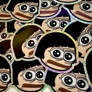 May include: A collection of stickers featuring a cartoon frog with black hair and large eyes. The stickers have a holographic finish and are arranged in a random pattern.