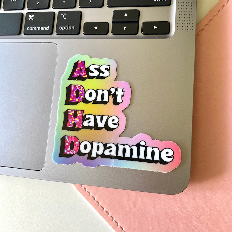 以下が含まれることがあります： 虹色のグラデーションの背景に、黒い文字で「Ass Don't Have Dopamine」と書かれたステッカー。ピンクのグリッターが散りばめられている。