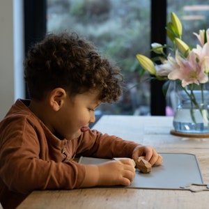 May include: A young child sits at a table playing with wooden geometric shapes on a grey silicone mat. The child is wearing a brown long-sleeved shirt.