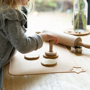 May include: A child is using a wooden cookie cutter to press a design into a cookie dough circle on a beige silicone baking mat. There are three other cookie dough circles on the mat. A wooden rolling pin and a metal star-shaped cookie cutter are also on the table.