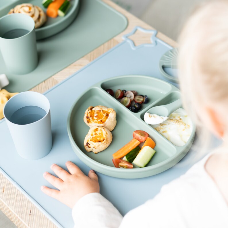 May include: A green silicone divided plate with a variety of foods for a child. The plate has sections for fruit, vegetables, and a small portion of yoghurt. There are two small pastries on the plate. The plate is on a blue silicone placemat.