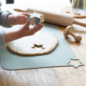 May include: A child uses a star-shaped cookie cutter to cut out a star from a round piece of cookie dough on a green silicone baking mat.