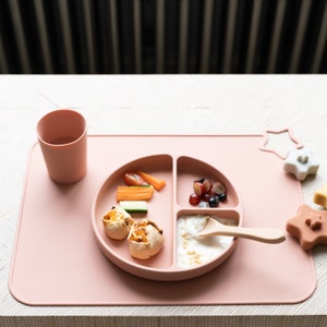 May include: A pink silicone divided plate with a spoon, a cup, and a variety of foods, including fruit, vegetables, and bread. The plate is on a pink silicone mat with a star-shaped toy.
