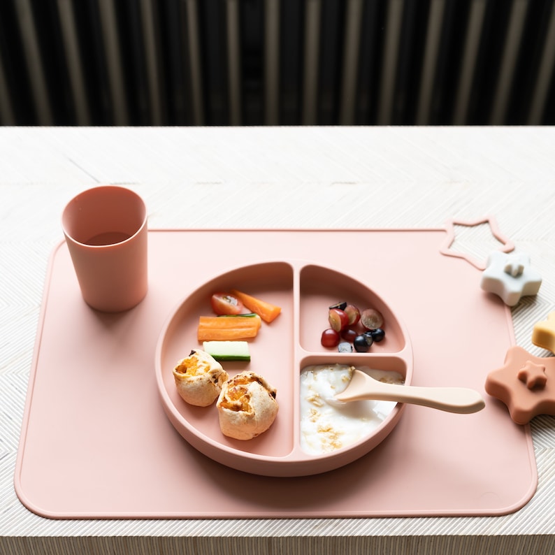 May include: A pink silicone divided plate with a spoon, a cup, and a variety of foods, including fruit, vegetables, and bread. The plate is on a pink silicone mat with a star-shaped toy.