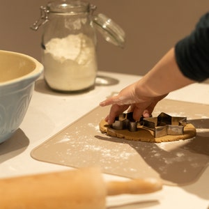 May include: A person is using cookie cutters to cut out star shapes from a piece of dough on a beige silicone baking mat. A rolling pin and a glass jar filled with flour are in the background.