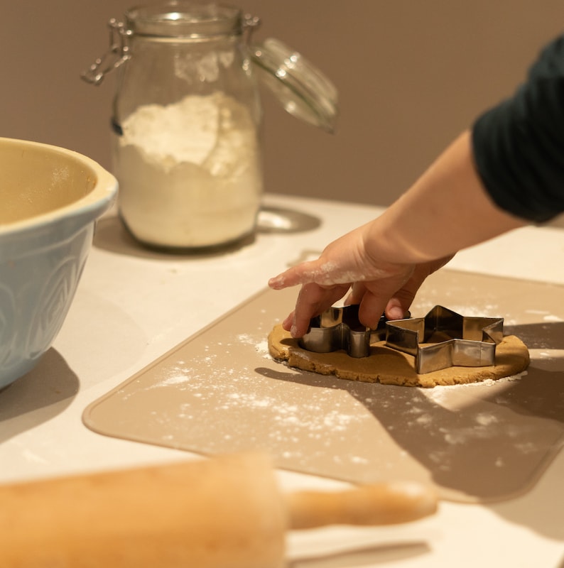 May include: A person is using cookie cutters to cut out star shapes from a piece of dough on a beige silicone baking mat. A rolling pin and a glass jar filled with flour are in the background.