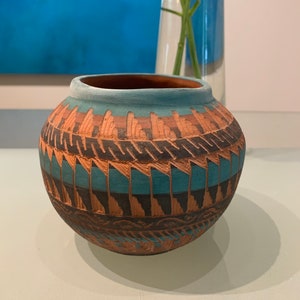 May include: A small, round, terracotta pot with a blue and brown geometric pattern. The pot is decorated with a repeating design of triangles and lines.