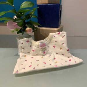 May include: Cream-colored baby onesie with a pattern of pink hearts and small flowers. The onesie has a round neckline with lace trim and a small bow. The brand name "Carter's" is visible on a tag.