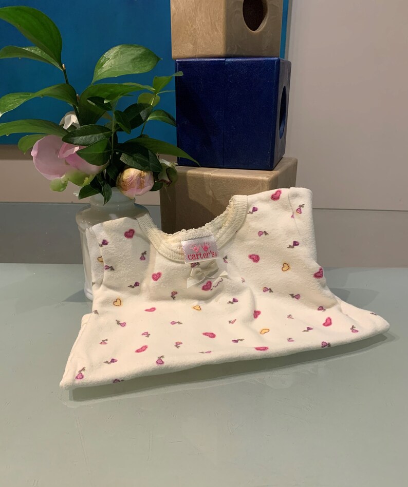 May include: Cream-colored baby onesie with a pattern of pink hearts and small flowers. The onesie has a round neckline with lace trim and a small bow. The brand name "Carter's" is visible on a tag.