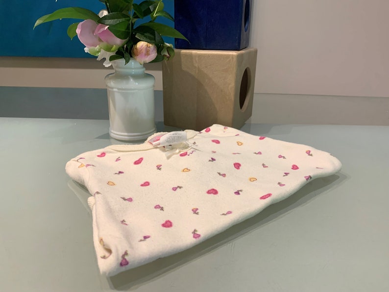 May include: Cream-colored baby onesie with a pattern of pink hearts and small purple and yellow shapes. The onesie is folded on a light blue surface. A white vase with pink flowers is in the background.