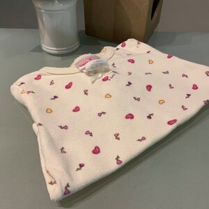 May include: A folded, off-white baby sleeper with a pattern of pink hearts and small purple flowers. The sleeper has a round neckline and a small bow. The fabric appears soft and fleecy.