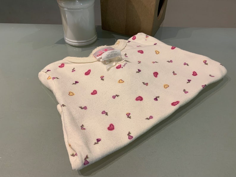 May include: A folded, off-white baby sleeper with a pattern of pink hearts and small purple flowers. The sleeper has a round neckline and a small bow. The fabric appears soft and fleecy.