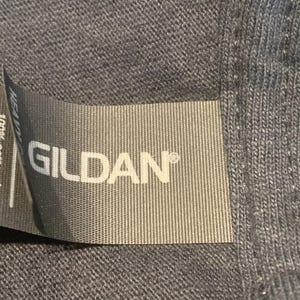 May include: Close-up of a gray Gildan brand tag sewn into a garment. The tag reads "100% cotton / algod&oacute;n / algod&atilde;o".