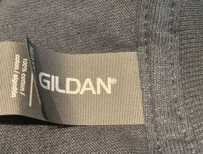 May include: Close-up of a gray Gildan brand tag sewn into a garment. The tag reads "100% cotton / algod&oacute;n / algod&atilde;o".