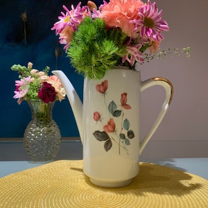 May include: A white ceramic pitcher with a gold handle and floral design, filled with pink and purple flowers, including carnations and daisies. The pitcher is sitting on a yellow woven placemat.