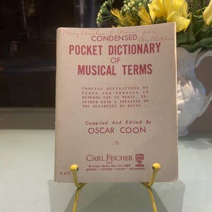 May include: A vintage "Pocket Dictionary of Musical Terms" book, with text in red and black. The cover reads "Merry Christmas to Alone - from Mrs. Chatelain". The book is propped up on a yellow metal stand.