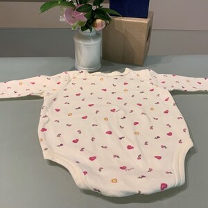 May include: A white baby bodysuit with long sleeves, covered in small pink hearts and yellow accents. The bodysuit has a round neckline and is made of soft fabric. The garment is designed for infants and toddlers.