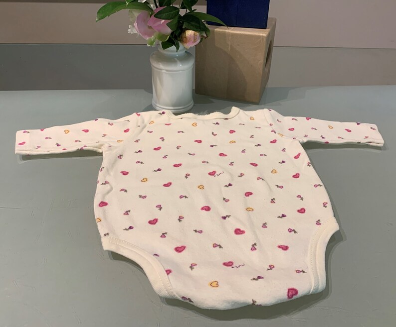 May include: A white baby bodysuit with long sleeves, covered in small pink hearts and yellow accents. The bodysuit has a round neckline and is made of soft fabric. The garment is designed for infants and toddlers.